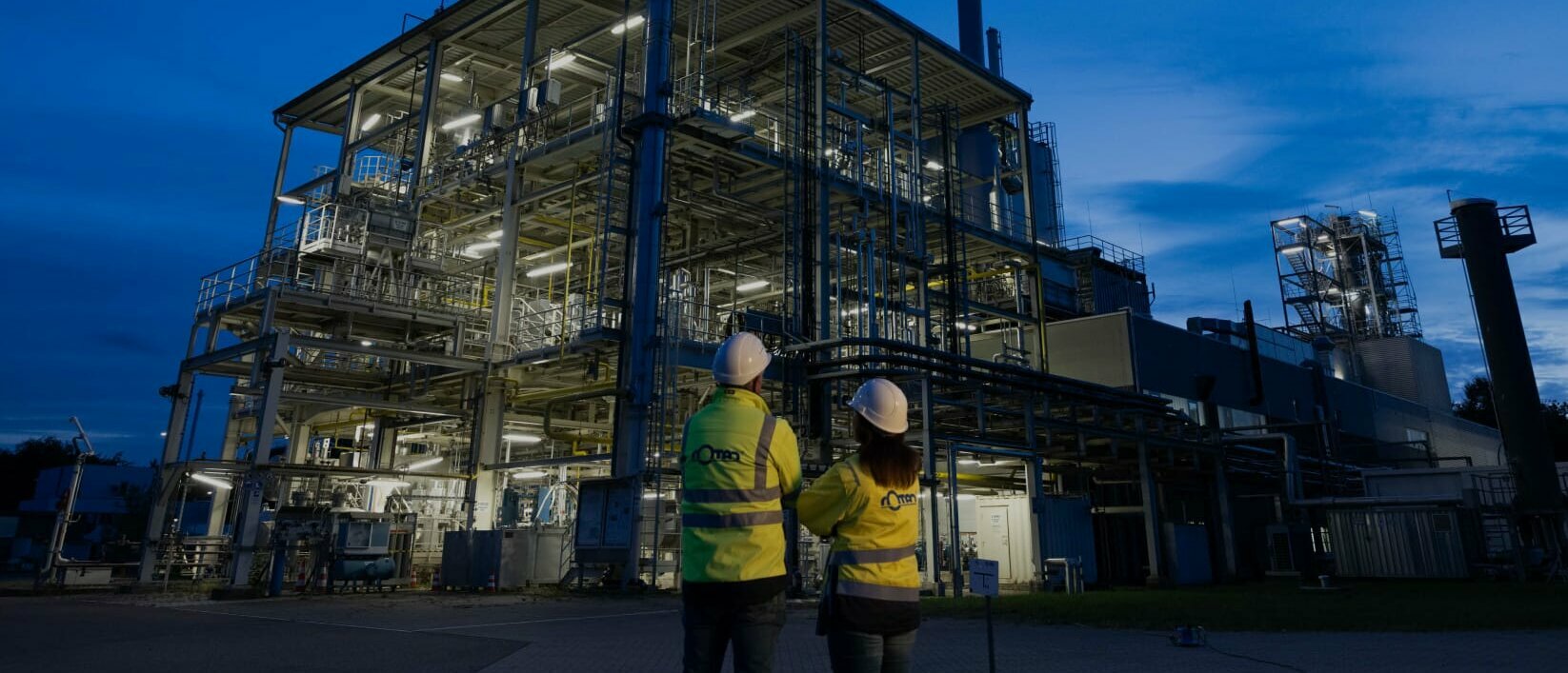 Zwei Mitarbeiter blicken auf eine beleuchtete Industrieanlage bei Nacht